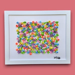 May include: A framed wall art piece featuring a dense, textured arrangement of small, colorful sculpted flowers in shades of pink, yellow, blue, and purple, interspersed with green leaves. The artwork is set in a white frame with a signature in the corner.
