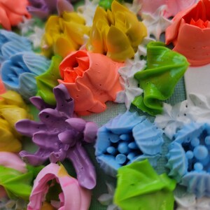 May include: A textured surface covered in thick, piped floral shapes made of colorful frosting. The arrangement features purple, yellow, light blue, pink, and orange flowers interspersed with bright green leaves and small white piped accents.