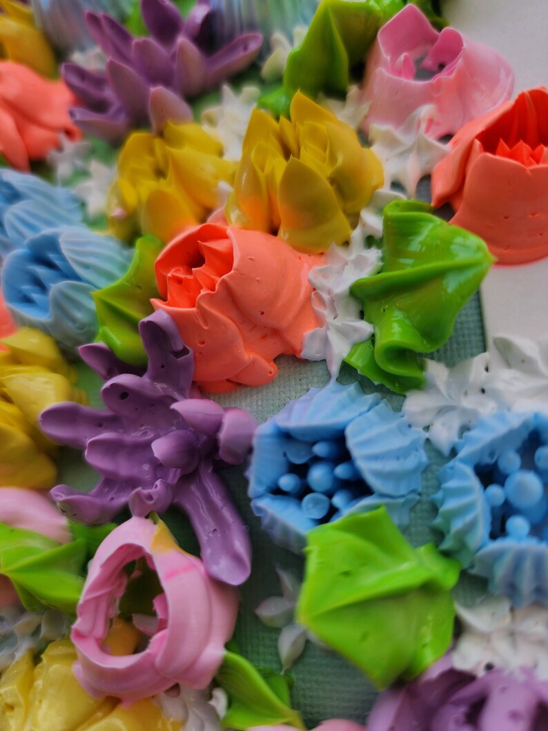 May include: A textured surface covered in thick, piped floral shapes made of colorful frosting. The arrangement features purple, yellow, light blue, pink, and orange flowers interspersed with bright green leaves and small white piped accents.