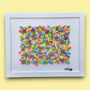 May include: A framed wall art piece featuring a dense, textured arrangement of small, multi-colored sculpted flowers in shades of pink, yellow, blue, purple, and green. The artwork is set against a white background within a white frame.