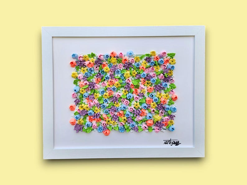 May include: A framed wall art piece featuring a dense, textured arrangement of small, multi-colored sculpted flowers in shades of pink, yellow, blue, purple, and green. The artwork is set against a white background within a white frame.