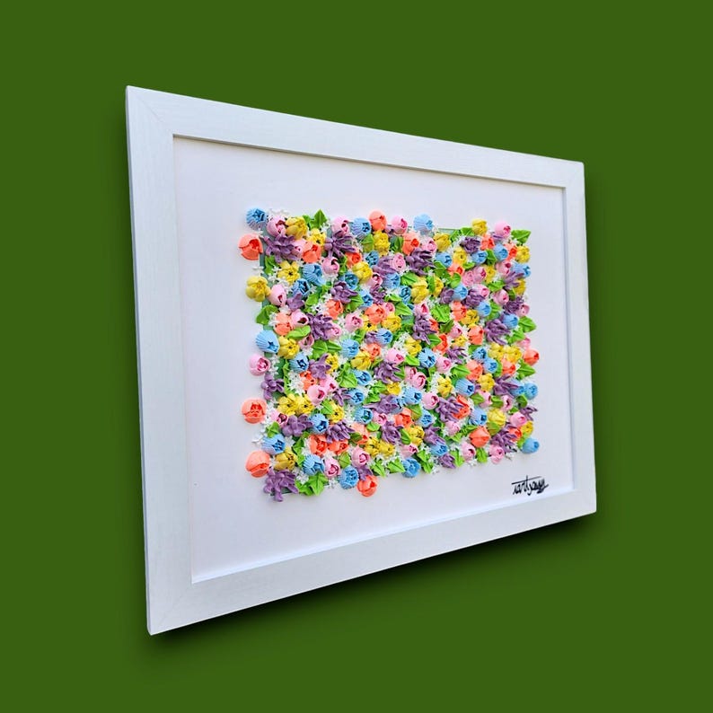 May include: A rectangular white frame containing a textured wall art piece featuring a dense arrangement of small, multi-colored floral shapes in shades of pink, yellow, blue, purple, and orange, with green leaves and a signature in the bottom corner.