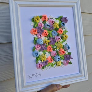 May include: A framed wall art piece featuring a textured, three-dimensional floral arrangement made of colorful piped paste. The composition includes pink, orange, yellow, blue, and purple flowers with green leaves on a white background. The text IX/XXV is visible.