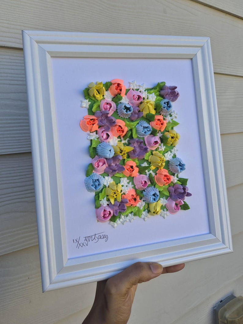 May include: A framed wall art piece featuring a textured, three-dimensional floral arrangement made of colorful piped paste. The composition includes pink, orange, yellow, blue, and purple flowers with green leaves on a white background. The text IX/XXV is visible.