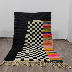 May include: A black and white checkered rug with a colorful stripe down the side. The rug has a fringe border.