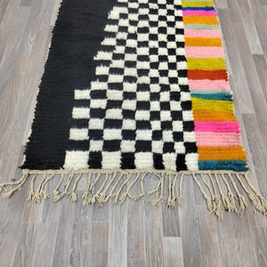 May include: A black and white checkered rug with a colorful striped border. The rug has a fringe border and is made of wool.