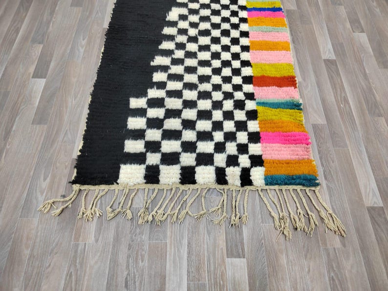 May include: A black and white checkered rug with a colorful striped border. The rug has a fringe border and is made of wool.