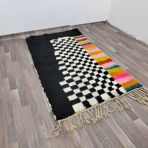 May include: A black and white checkered rug with a colorful border. The border features stripes of pink, yellow, orange, and blue. The rug has fringe on all sides.