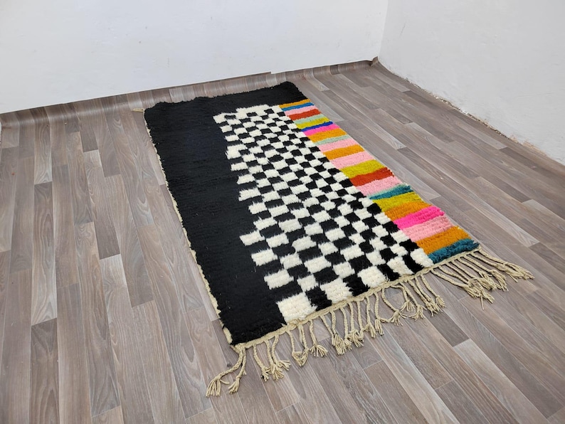 May include: A black and white checkered rug with a colorful border. The border features stripes of pink, yellow, orange, and blue. The rug has fringe on all sides.