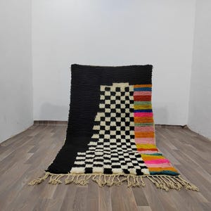 May include: A black and white checkered rug with a colorful stripe down the side. The rug has a fringe border.
