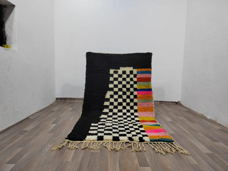May include: A black and white checkered rug with a colorful stripe down the side. The rug has a fringe border.