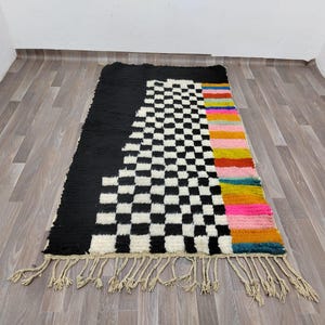May include: A black and white checkered rug with a colorful stripe down the side. The rug has a fringe border.