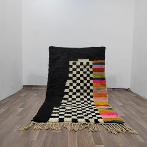 May include: A black and white checkered rug with a colorful stripe down the side. The rug has a fringe border.