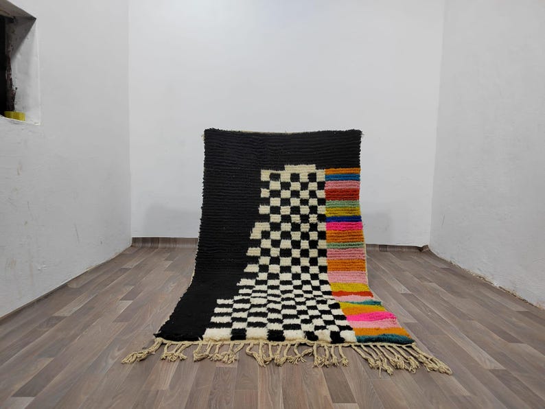 May include: A black and white checkered rug with a colorful stripe down the side. The rug has a fringe border.
