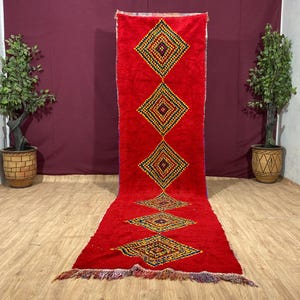 May include: A red Moroccan rug with a geometric pattern of yellow, blue, and green diamonds. The rug has a fringe border and is laid out on a wooden floor.