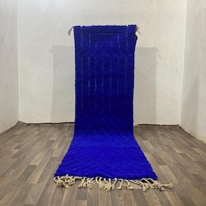 Puede incluir: Una alfombra marroquí larga y azul con un patrón geométrico y flecos. La alfombra está colgada en una pared blanca.