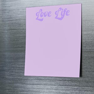 May include: A lilac sticky note with the words "Love Life" in a cursive font is attached to a stainless steel fridge door with magnetic circles. The note is blank and ready for writing.