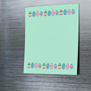 Può includere: Un post-it verde chiaro con un bordo di colorati disegni di uova di Pasqua è attaccato alla porta di un frigorifero in acciaio inossidabile con magneti. Il biglietto è vuoto, pronto per essere scritto.