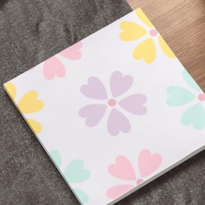 May include: A square notepad featuring a floral pattern in pastel shades of yellow, pink, purple, and teal. The notepad is on a dark grey surface, near a laptop and a plant pot.