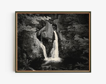 Könnte beinhalten: Ein Schwarzweißfoto eines Wasserfalls, der eine felsige Klippe in einem üppigen Wald hinunterstürzt. Das Wasser fließt über einen großen Felsen in der Mitte des Bildes und erzeugt einen dramatischen Effekt.