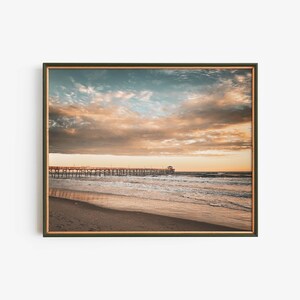 May include: A wooden pier stretches out into the ocean, with a cloudy sky in shades of pink, orange, and blue. The water is a light blue, with white waves breaking on the shore. The beach is sandy and deserted.