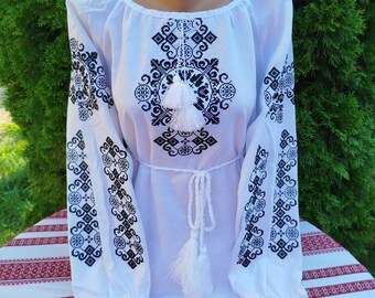 May include: White embroidered blouse with black geometric patterns and a white tassel. The blouse has a round neckline and long sleeves.