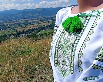 May include: White embroidered blouse with green and black geometric patterns. The blouse features a green tassel and is set against a backdrop of a mountain landscape under a blue sky. The sleeves are long and the bottom hem is red.