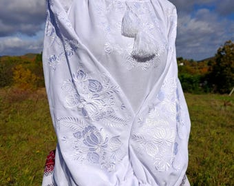 May include: White embroidered blouse with long sleeves, adorned with floral patterns and a tassel. A red beaded necklace complements the blouse, which is paired with a skirt featuring a red and white geometric design. The sleeves are gathered at the wrist.