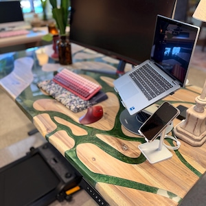 May include: A wooden desk with a live edge design, featuring a river of green epoxy resin. A laptop, monitor, and phone are on the desk. A treadmill is visible below the desk. The desk has a modern, natural aesthetic.