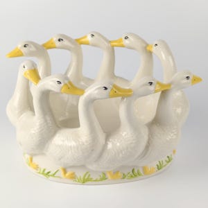 Puede incluir: Un centro de mesa de cerámica blanca con un anillo de gansos. Cada ganso tiene un pico amarillo y está dispuesto alrededor del borde del cuenco. La base del cuenco tiene un diseño de hierba verde y amarilla.