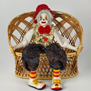 May include: A clown doll seated in a woven wicker chair. The doll has a red hat, gold vest, patterned trousers, and oversized shoes. The clown's painted face features a red bow tie and a red button on the vest.