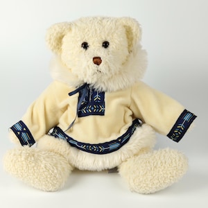 Puede incluir: Un oso de peluche crema con un abrigo amarillo claro con patrones geométricos azules y blancos. El oso tiene una nariz marrón y ojos negros. El abrigo tiene adornos azules y blancos. El oso está sentado sobre un fondo blanco.