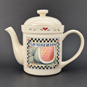 Puede incluir: Tetera de cerámica color crema con tapa, boquilla y asa. La tetera presenta un diseño cuadrado con una rodaja de sandía y la palabra "WATERMELON". La tapa tiene un pequeño pomo y está decorada con corazones y hojas.