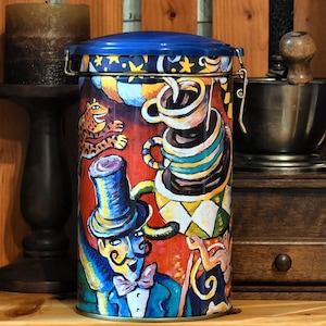 May include: A cylindrical tin with a blue lid and a clasp. The tin is decorated with a colourful circus-themed design featuring a magician, coffee cups, and a leopard. The background is red.