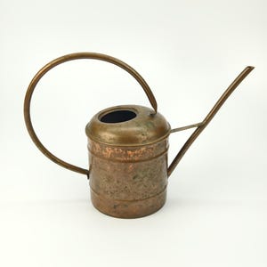 Può includere: Un annaffiatoio vintage in rame con manico curvo e lungo beccuccio. Il contenitore ha un corpo cilindrico e una parte superiore rotonda con un foro di riempimento. Il rame ha una patina invecchiata, che gli conferisce un aspetto rustico. Perfetto per il giardinaggio.