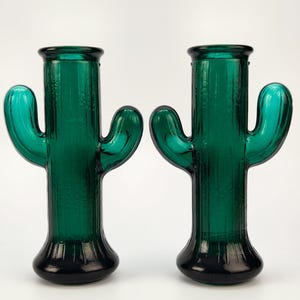 Peut inclure: Deux bougeoirs en verre vert en forme de cactus. Les bougeoirs ont une base vert foncé et un corps vert clair avec deux bras incurvés. Les bougeoirs sont en verre et ont une surface texturée.