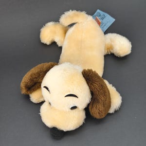 Könnte beinhalten: Ein Stoffhund-Spielzeug in liegender Position. Der Körper ist hellbraun, die Ohren braun und die Nase schwarz. Die Augen sind mit schwarzem Faden geschlossen. Ein blaues Etikett ist befestigt.