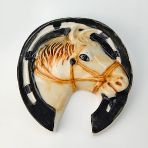 Puede incluir: Plato de cerámica en forma de herradura con cabeza de caballo. La cabeza del caballo es de color crema con detalles marrones, incluyendo la crin y la brida. La herradura es negra y blanca, creando un borde contrastante. Un artículo decorativo de temática ecuestre.