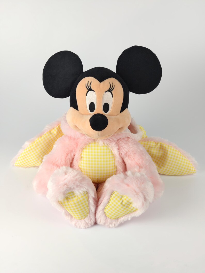 May include: A plush Minnie Mouse doll wearing a pink bunny costume with yellow gingham accents. The doll has black ears, a pink face, and a black nose. The bunny costume features long ears and fluffy pink fur.