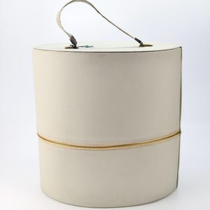 Puede incluir: Sombrerera cilíndrica de color crema con cremallera dorada y asa curva. La caja está hecha de un material texturizado y tiene la parte superior e inferior redondeadas. El asa está sujeta con herrajes metálicos.