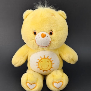 Könnte beinhalten: Ein gelber Plüschbär mit einem lächelnden Sonnenemblem auf dem Bauch. Der Bär hat eine weiße Schnauze, eine orangefarbene Nase und gestickte Augen. Seine Pfoten weisen herzförmige Designs auf. Das Spielzeug befindet sich vor einem dunklen Hintergrund.