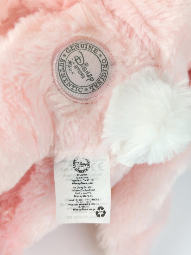 May include: Close-up of a pink plush toy with a white pom-pom and a Disney Store label. The label features the Disney Store logo and text. The plush toy has a soft, fluffy texture.