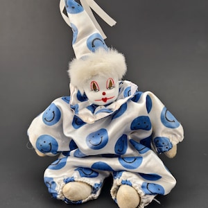 May include: A vintage clown doll with a white and blue smiley face pattern. The doll has a white fur collar, a pointed hat, and a painted face with red lips and rosy cheeks. The clown's outfit includes a matching hat and pants.