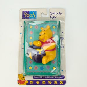 May include: A Winnie the Pooh Switch-Ups wall decor in a plastic package. The yellow Pooh figure is holding a pot of honey on a green background with a patterned design. The packaging includes the text "Pooh" and "Switch-Ups".