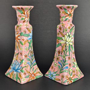 Puede incluir: Un par de candelabros de cerámica con un color base rosa y un patrón floral pintado a mano. El diseño incluye varias flores en tonos naranja, amarillo y azul, acentuadas con hojas verdes. Cada candelabro tiene una parte superior y base acampanadas.