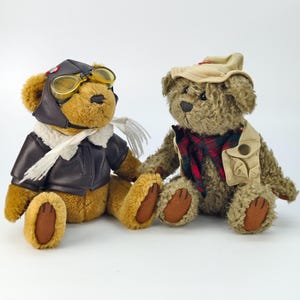Puede incluir: Dos osos de peluche vestidos con trajes temáticos. Uno lleva una chaqueta de aviador de cuero marrón, gafas y una bufanda blanca. El otro oso viste un chaleco color canela, una camisa a cuadros y un sombrero. Ambos osos tienen patas marrones.