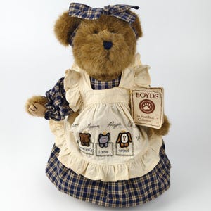 May include: A brown plush bear wearing a blue and cream plaid dress and a matching bow. The bear has a cream apron with embroidered animal designs and the words "Little Angels". A tag reads "Boyds The Head Bean Collection."