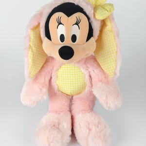 May include: A plush Minnie Mouse doll dressed in a pink bunny costume. The costume features yellow gingham accents on the ears, bow, and belly. Minnie's face is black, with large eyes and a black nose.