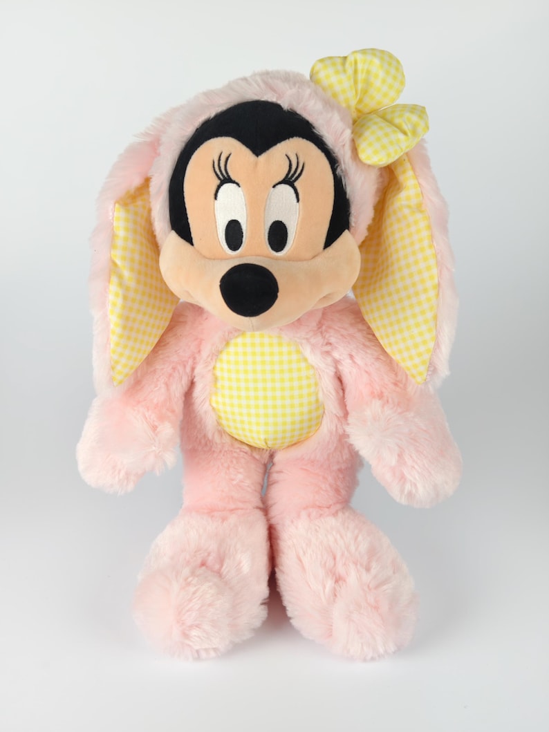 May include: A plush Minnie Mouse doll dressed in a pink bunny costume. The costume features yellow gingham accents on the ears, bow, and belly. Minnie's face is black, with large eyes and a black nose.