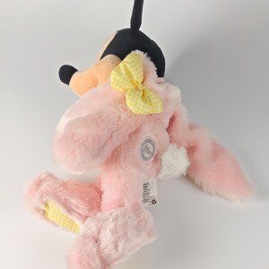 May include: A plush toy featuring a pink bunny costume with a yellow gingham bow and ears. The toy has a black and tan head, resembling a cartoon character. The bunny's feet and ears have yellow gingham accents. A white tag is attached.